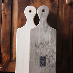 May include: Two wooden cutting boards, one is natural wood and the other is stained dark brown. The dark brown board has a triple moon symbol carved into it.