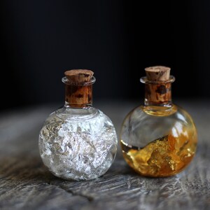 May include: Two small glass bottles with cork stoppers. One bottle contains silver flakes and the other contains gold flakes.