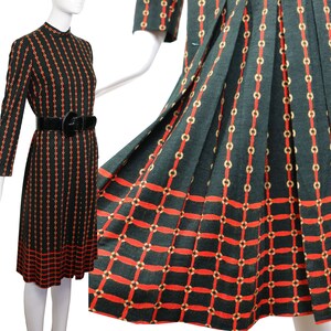 Op de afbeelding: Een vintage zwarte jurk met een rood en goud kettingpatroon. De jurk heeft lange mouwen, een hoge halslijn en een zwarte riem. De rok is geplooid. De jurk wordt op een mannequin getoond.