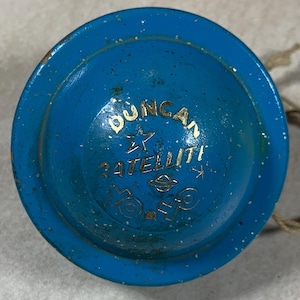 Könnte beinhalten: Ein Vintage-Yo-Yo von Duncan Satellite in Blau mit goldfarbenen Buchstaben. Das Yo-Yo hat eine Schnur und weist Gebrauchsspuren auf. Der Text auf dem Yo-Yo lautet "Duncan Satellite".