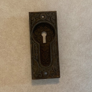May include: A vintage, ornate, cast iron keyhole cover with a detailed floral design. The cover is dark brown and has a keyhole in the center.