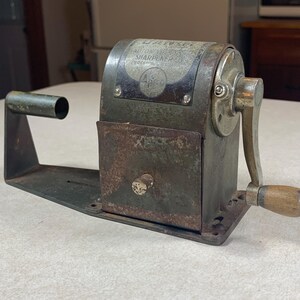 May include: A vintage metal pencil sharpener with a weathered dark gray finish, featuring a hand crank with a wooden handle, a rectangular base, and a metal plate on the top with the text "Dandy Automatic Sharpener" clearly visible.