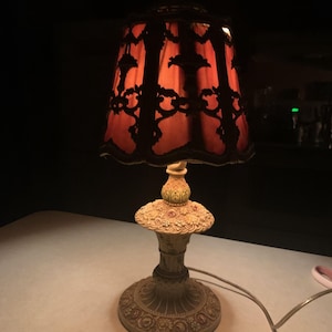 May include: An antique table lamp with a red lampshade featuring a black floral design. The lamp has a detailed, ornate base with a cream and gold finish. The lamp is on a white surface.