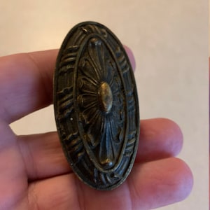 May include: An oval-shaped, antique brass door knob with a floral design. The knob has a raised center with a circular, brass-colored accent.