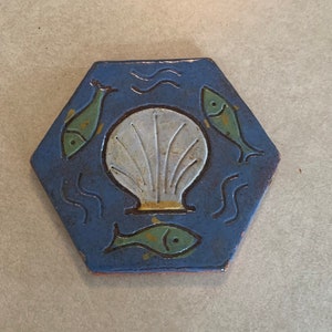 May include: A blue hexagonal ceramic tile with a white seashell and three green and yellow fish. The fish are swimming in blue wavy lines.