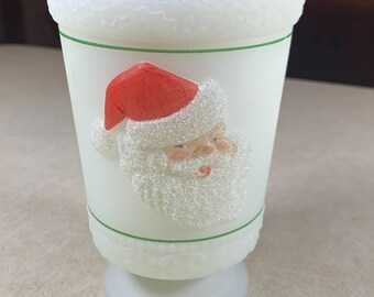 May include: A white glass Christmas goblet featuring a Santa Claus design. The goblet has a scalloped rim, a green band, and a pedestal base. The Santa Claus image has a red hat and a white beard.