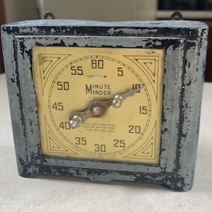 May include: An antique Minute Minder timer with a square, distressed metal frame. The clock face is cream-colored with black numerals and hands. The text "MINUTE MINDER" is centered on the face. The timer is likely from the mid-20th century.