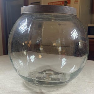May include: A large, clear glass jar with a round, ribbed body and a dark metal lid. The jar sits on a white surface, reflecting the surrounding environment. The jar is likely for storage or display.