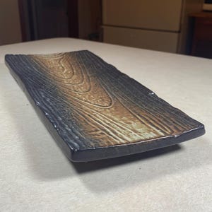 May include: A rectangular ceramic serving platter with a wood grain design. The platter has a dark brown border and a lighter brown and tan center, resembling the look of a wooden plank. The surface has a textured appearance.