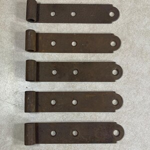 May include: Five rectangular metal strap hinges arranged in a vertical column. Each hinge features a dark brown, weathered finish with a cylindrical barrel at one end and three circular mounting holes along the flat, elongated metal plate.