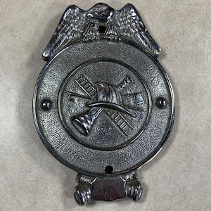 May include: A circular silver-toned metal badge featuring an eagle at the top. The center displays a relief design of a firefighter helmet, a ladder, and a tool. The badge has a textured finish and small holes for mounting on a flat surface.