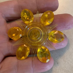 May include: A yellow plastic sun-shaped brooch with six oval-shaped gems. The brooch has a starburst design with points radiating from the center.