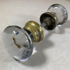 May include: A door knob set featuring two clear, spherical glass knobs connected by a central metal spindle. Each glass knob is mounted on a circular, tarnished brass-colored metal base, showing signs of age and wear on the metallic components.