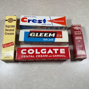 May include: A collection of vintage toothpaste boxes, including brands like Squibb, Crest, Gleem, Colgate, and Elizabeth Arden. The boxes are in various colors, with text and logos from the mid-20th century. The Squibb box is yellow, while the Colgate box is red.