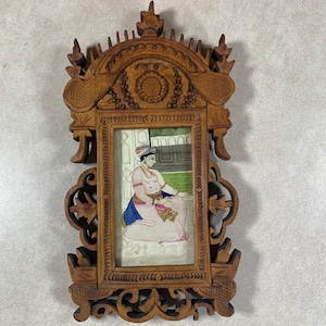 May include: A rectangular wall hanging featuring a hand-painted portrait of a seated figure in traditional attire, enclosed within an ornate, carved brown wooden frame with intricate floral and geometric patterns and a decorative arched top.