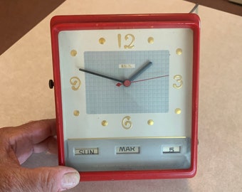 Könnte beinhalten: Eine rote Vintage-Uhr mit weißem Zifferblatt und goldenen Akzenten. Die Uhr verfügt über eine Kalenderfunktion mit den Wochentagen und dem Datum. Die Uhr ist auf Sonntag, den 5. März eingestellt.