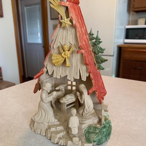 May include: A vintage Christmas nativity scene made of plastic. The scene depicts the birth of Jesus in a stable with Mary, Joseph, and a shepherd. The stable is decorated with a red roof and a green tree. A star shines above the stable.
