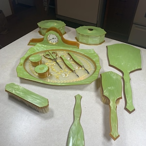 May include: A vintage vanity set in light green pearlescent material with brown trim. The set includes a clock, a tray with scissors and tools, two lidded jars, a tissue box, a powder box, a hand mirror, a hairbrush, and a clothes brush.