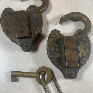 May include: Two antique heart-shaped padlocks with open shackles and a brass key. The locks have a dark, aged appearance with visible wear and tear. The key is made of brass and is attached to a metal ring.