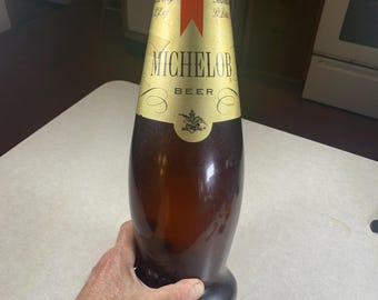 May include: A large, brown glass Michelob beer bottle with a gold and red label. The label features the brand name and the word "BEER". The bottle has a unique shape and is held by a person.