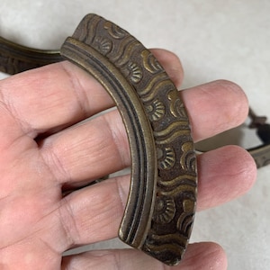 May include: A pair of antique brass drawer pulls with an ornate design. The pulls are shaped like a half-circle and have a textured surface.