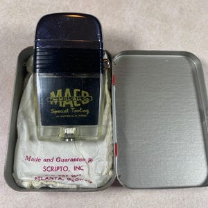 May include: A vintage black and clear lighter with the text "MACD The MILL-ALL Co." and "Special Tooling" in a silver metal tin. The lighter is in a white paper wrapping with the text "Made and Guaranteed by SCRIPTO, INC. ATLANTA, GEORGIA."