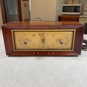 May include: Vintage Philco clock radio with a wooden frame and a cream-colored face. The clock features a manual dial, a start dial, and the brand name "PHILCO" printed at the bottom. The clock's face displays the time with black numerals.