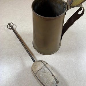 May include: An antique brass mug with a hinged lid and a handle. A long, twisted metal tool with a stone-like head rests beside the mug. The mug and tool appear aged, suggesting vintage or historical use.