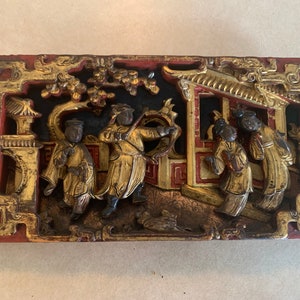 May include: A carved wooden panel depicting a scene from Chinese mythology. The panel is painted in gold, red, and black, and features figures in elaborate costumes. The panel is a traditional Chinese art form.