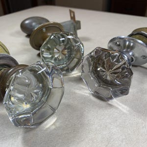 May include: A collection of three vintage door knobs featuring clear, faceted glass handles with an octagonal shape. Each knob is attached to a circular metal base plate, with finishes including aged brass and polished chrome hardware components.