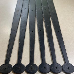 May include: Six long, black metal straps with a hammered texture along the edges. Each strap features a circular end with a centered hole and additional holes spaced along the length of the tapered body for mounting purposes.