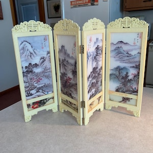 May include: A four-panel room divider made of cream-colored plastic with a traditional Chinese design. Each panel features a different landscape scene with mountains, trees, and water. The panels are hinged together and can be folded for storage.