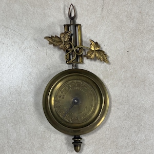 May include: A brass clock pendulum featuring a circular dial with engraved numbers and the text W.L. GILBERT CLOCK CO. The pendulum has a decorative leaf motif at the top, a central needle, and a pointed finial at the bottom, all in a metallic finish.