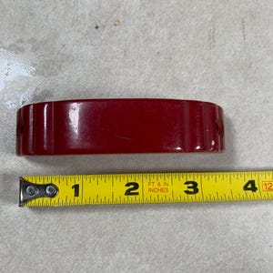 May include: A rectangular, glossy, red object with rounded edges, approximately 3.5 inches long. Two small holes are visible on the sides. A yellow measuring tape with black markings is placed next to the object.