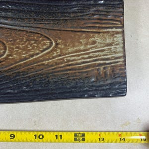 K&ouml;nnte beinhalten: Rechteckige Servierplatte aus Keramik mit Holzmaserung. Die Platte hat eine dunkelbraune und beige Farbgebung mit einer strukturierten Oberfl&auml;che. Die Platte ist etwa 30 cm lang.
