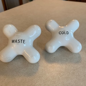 May include: Two white ceramic faucet handles with a cross-shaped design. One handle is labeled "WASTE" and the other is labeled "COLD" in black text. The handles have a slightly aged appearance.