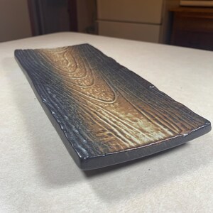 K&ouml;nnte beinhalten: Rechteckige Servierplatte aus Keramik mit Holzmaserungsdesign. Die Platte hat einen dunkelgrauen Rand und eine Mitte mit Braun- und Goldt&ouml;nen, die nat&uuml;rlichem Holz &auml;hneln. Die Kanten sind leicht uneben, was ihr ein rustikales Aussehen verleiht.