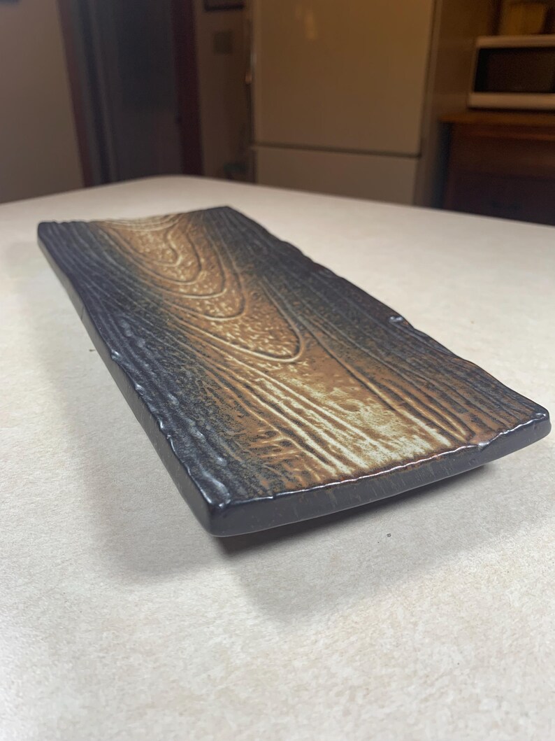 K&ouml;nnte beinhalten: Rechteckige Servierplatte aus Keramik mit Holzmaserungsdesign. Die Platte hat einen dunkelgrauen Rand und eine Mitte mit Braun- und Goldt&ouml;nen, die nat&uuml;rlichem Holz &auml;hneln. Die Kanten sind leicht uneben, was ihr ein rustikales Aussehen verleiht.