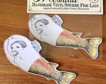 May include: Two handmade vinyl stickers of a mermaid with a rainbow trout tail. The stickers are on a wooden surface. The text on the sticker says "She's 50% Rainbow Trout, 100% Attitude Mermaid Handmade Vinyl Sticker Fish Lady original artwork by Rosie Ferne Illustration"