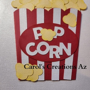 May include: Red and white striped popcorn box with the word "POPCORN" in white letters. The box is filled with yellow popcorn pieces. The bottom of the box has the text "Carol's Creations Az".