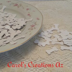 May include: A white ceramic plate filled with white paper bone shapes. The plate is on a white lace tablecloth. The bone shapes are scattered on the tablecloth.