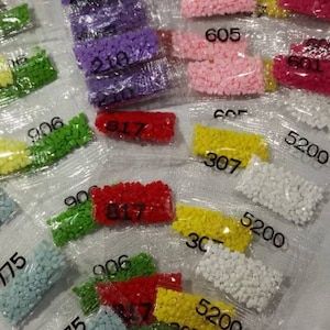 May include: Close-up of small plastic bags filled with colorful beads. The bags are labeled with numbers, including 727, 775, 906, 817, 605, 601, 307, and 5200.