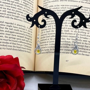May include: A pair of silver hoop earrings with a yellow glass bead charm. The earrings are on a black stand in front of an open book.