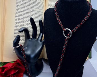 Può includere: Una collana a catena nera e rossa con un ciondolo a forma di anello d'argento. La catena è composta da piccoli anelli di metallo collegati tra loro.