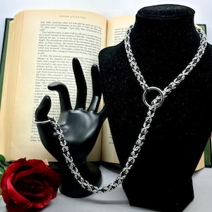 May include: A silver and black chainmail necklace with a large silver ring. The necklace is displayed on a black mannequin bust. The necklace is on a table with a book and a red rose.