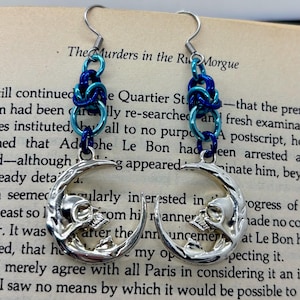 May include: Silver-toned earrings featuring a crescent moon design with a skull inside. The earrings have blue and teal chainmaille accents and are suspended from silver hooks. The background is a page from a book.