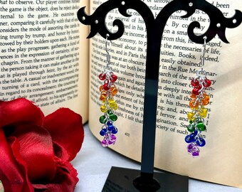 May include: Rainbow colored beaded earrings with silver chain links. The earrings are displayed on a black stand against a book.