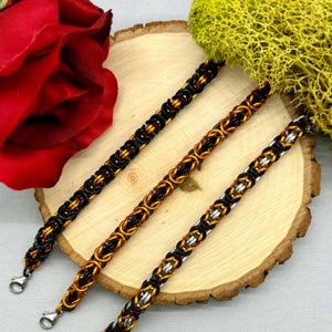 May include: Three chain bracelets in black, gold, and silver with a black and gold pattern. The bracelets are displayed on a wooden surface.
