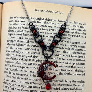 May include: A silver chain necklace with black and red accents. The pendant is a red crescent moon with a wolf or dog silhouette and a red teardrop gem. The necklace is displayed on an open book.