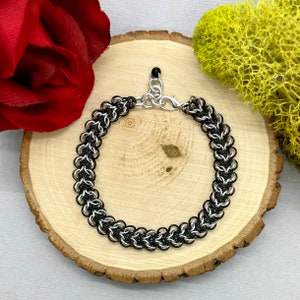 May include: A handcrafted bracelet featuring a chainmail design with alternating black and silver rings. The bracelet is displayed on a wooden slice, with a red rose and green moss in the background. The bracelet has a silver-colored clasp.